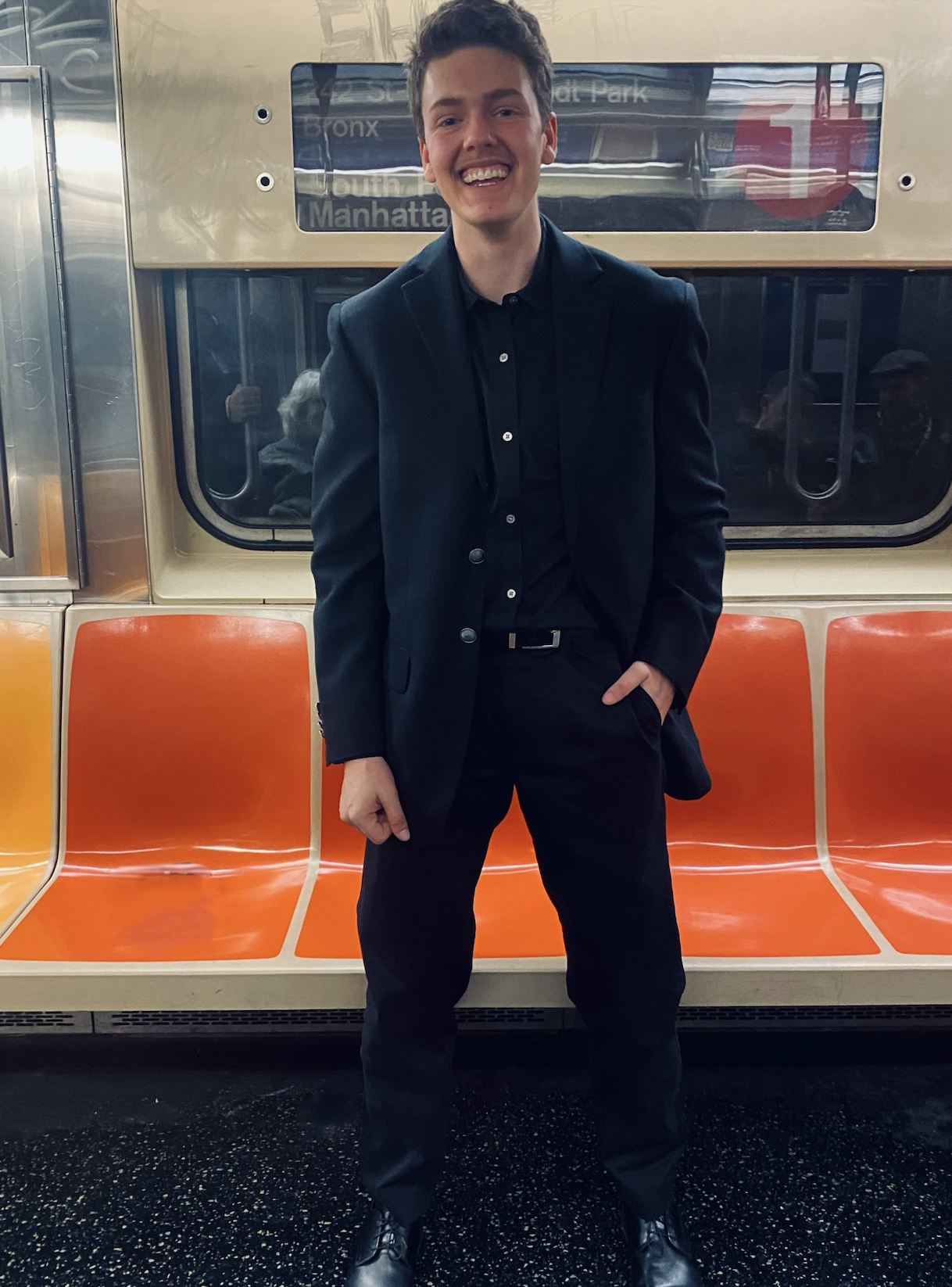 A student, dressed in a black suit, standing in a subway car, smiling